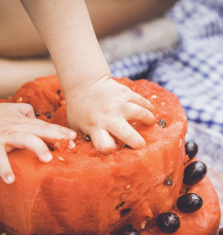 Beikostberatung.jpg Eine kleine Hand greift nach einer frischen watermelon mit schwarzen Kernen.