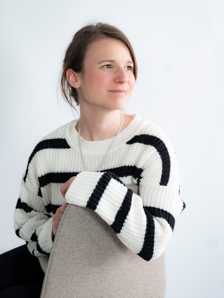 Laura.jpg Eine Person mit einem gestreiften Pullover sitzt auf einem Stuhl und blickt nachdenklich zur Seite.